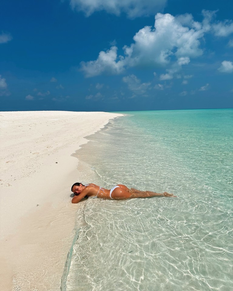 Woman in white bikini lying in shallow clear water on a white sand beach.