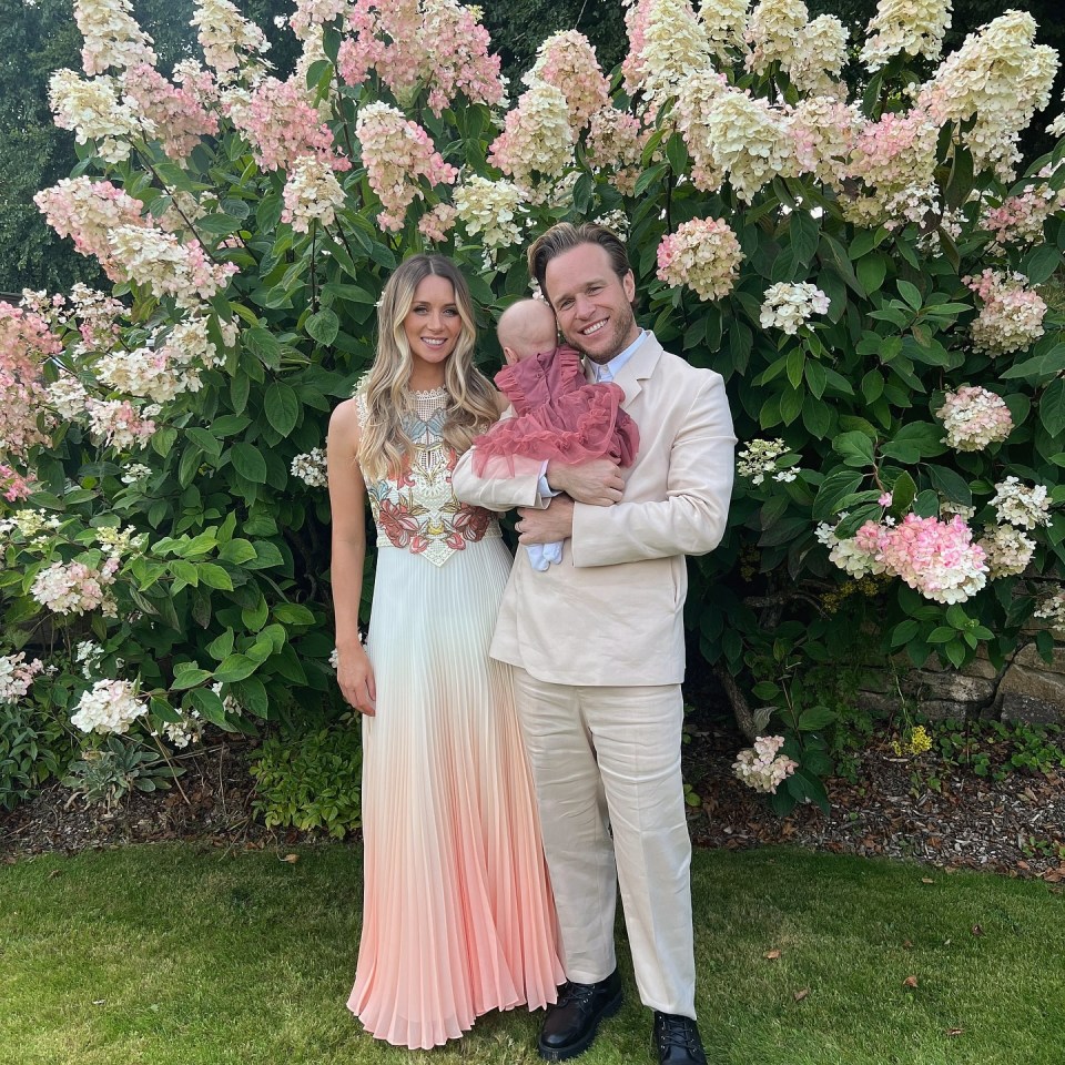 Family photo of a man and woman in wedding attire holding their baby in front of hydrangeas.