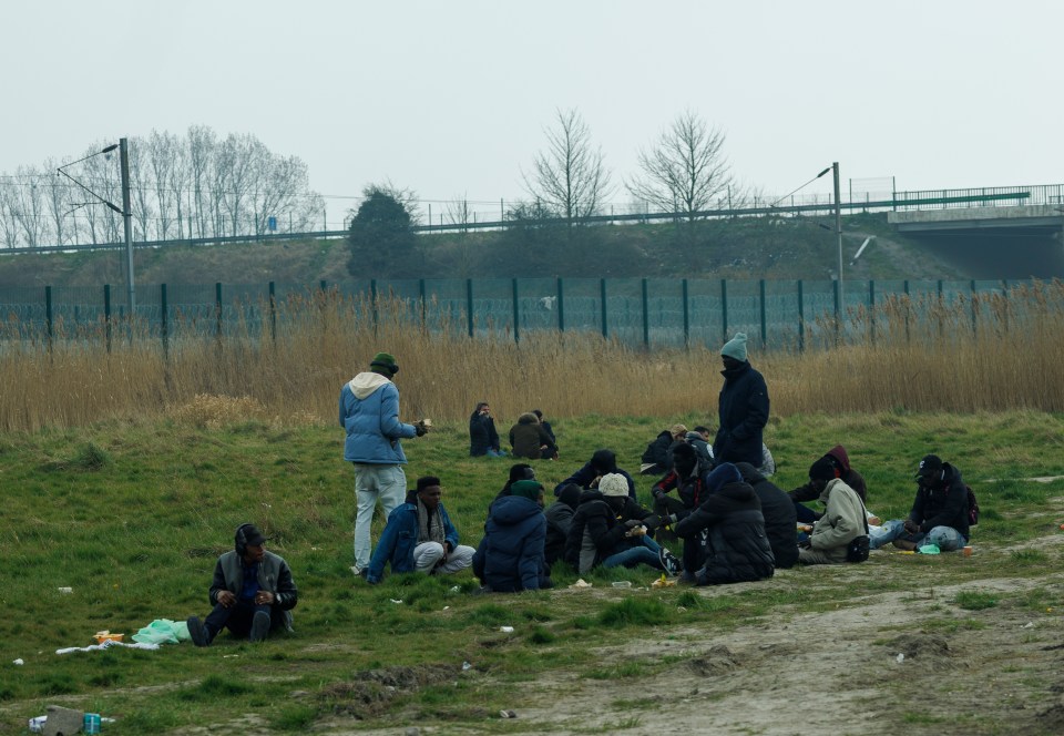 Migrants sitting on the ground near a fence, receiving food.