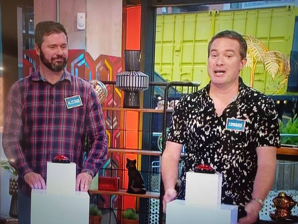 Two game show contestants at podiums with buzzers.