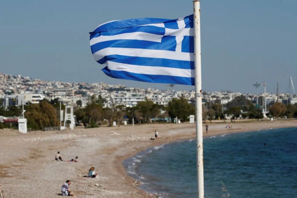An image collage containing 1 images, Image 1 shows Greek flag on a beach with people in the background