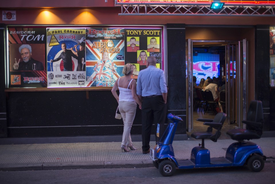Pedestrians view posters advertising British entertainers outside a venue.