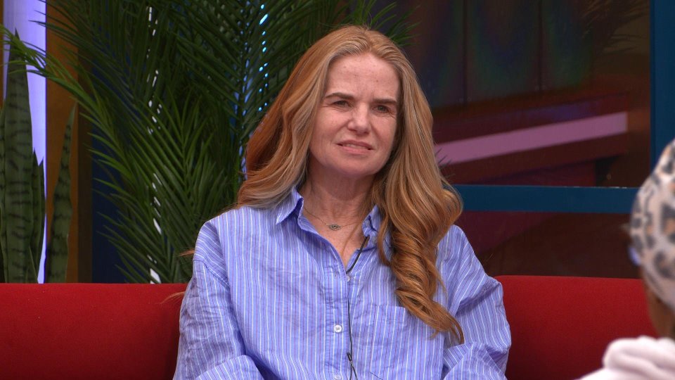 A woman with long, light brown hair sits on a red couch and speaks.