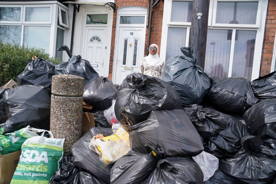 A woman stands in front of a large pile of garbage bags on a residential street.
