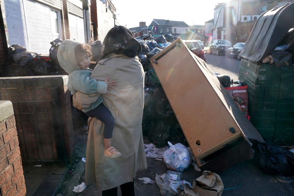 A woman carries a young child past overflowing rubbish on a residential street.