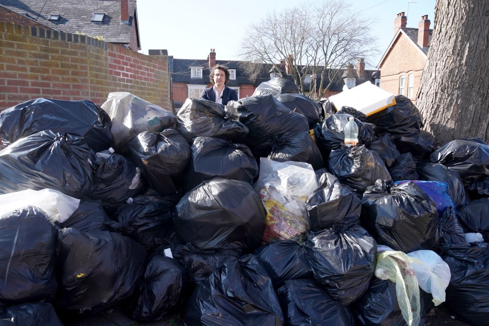 A man stands behind a large pile of black garbage bags overflowing onto a street.