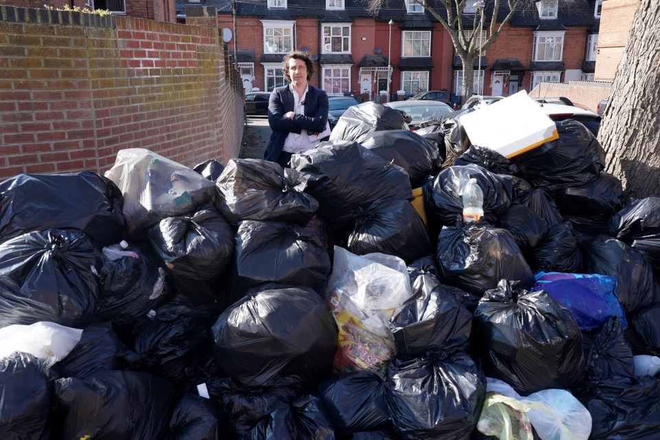A man stands amidst a large pile of overflowing black garbage bags on a residential street.