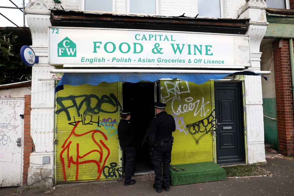 Two police officers standing outside Capital Food & Wine, a shop with graffiti on its shutters.