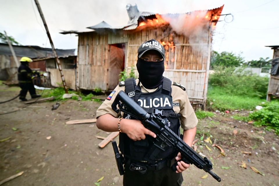 Police officer with rifle standing guard near a burning building.