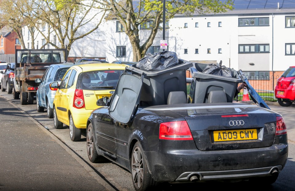 Cars queuing at a mobile household waste collection point with overflowing bins.
