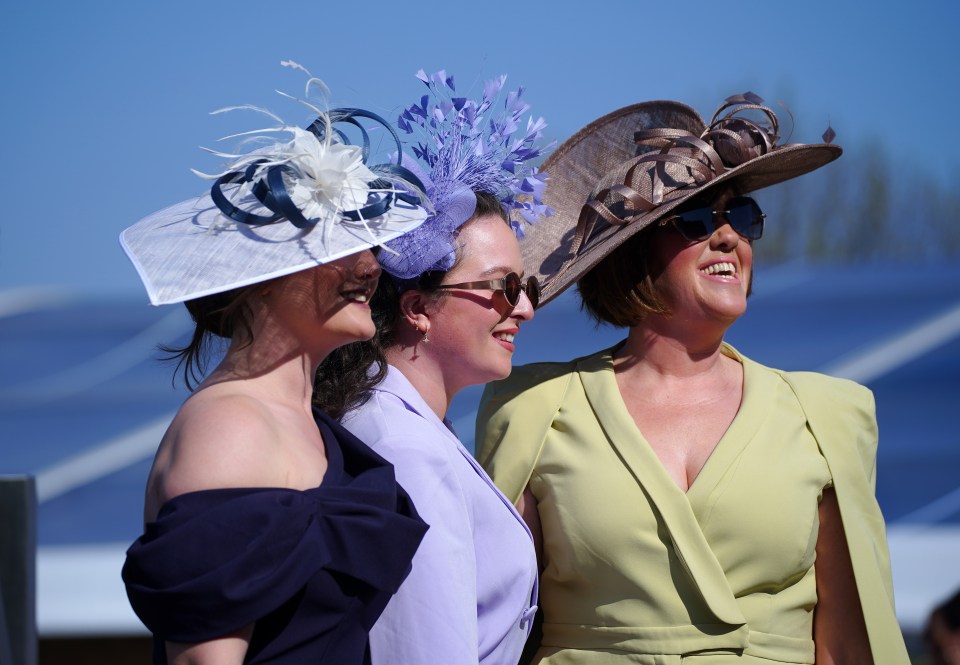 Three women wearing stylish hats and sunglasses at a horse race.