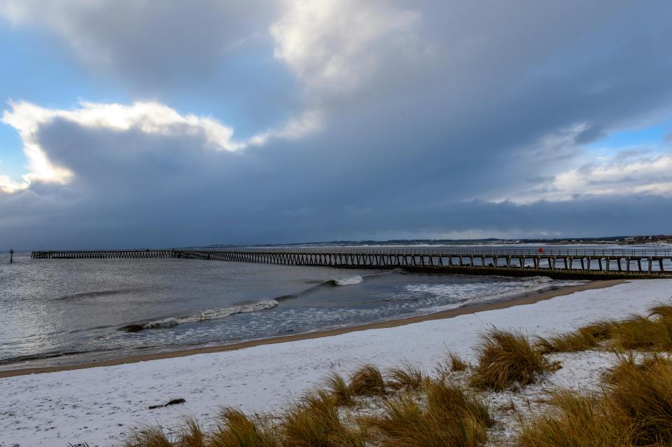 Snowy beach with pier and ocean under a cloudy sky.