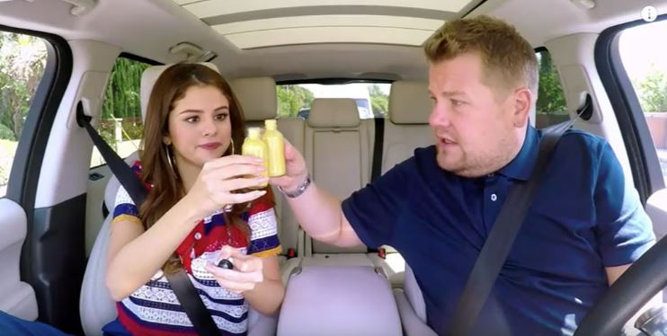 Selena Gomez and James Corden in a car, holding small bottles of yellow liquid.