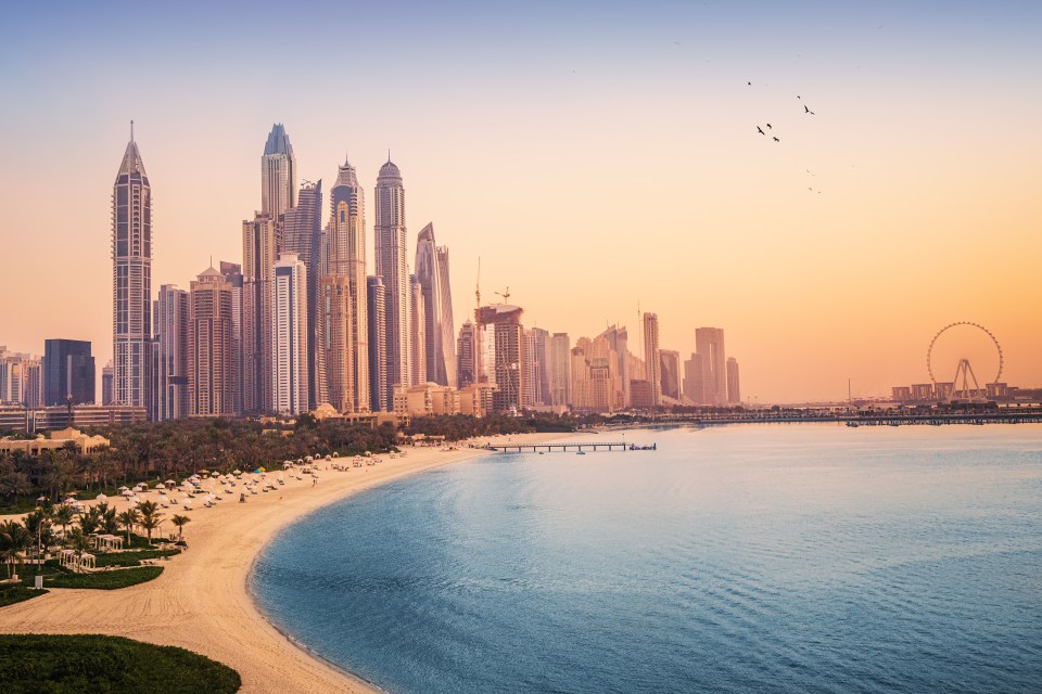 Sunset view of Dubai Marina and JBR, featuring skyscrapers, a Ferris wheel, and a sandy beach.