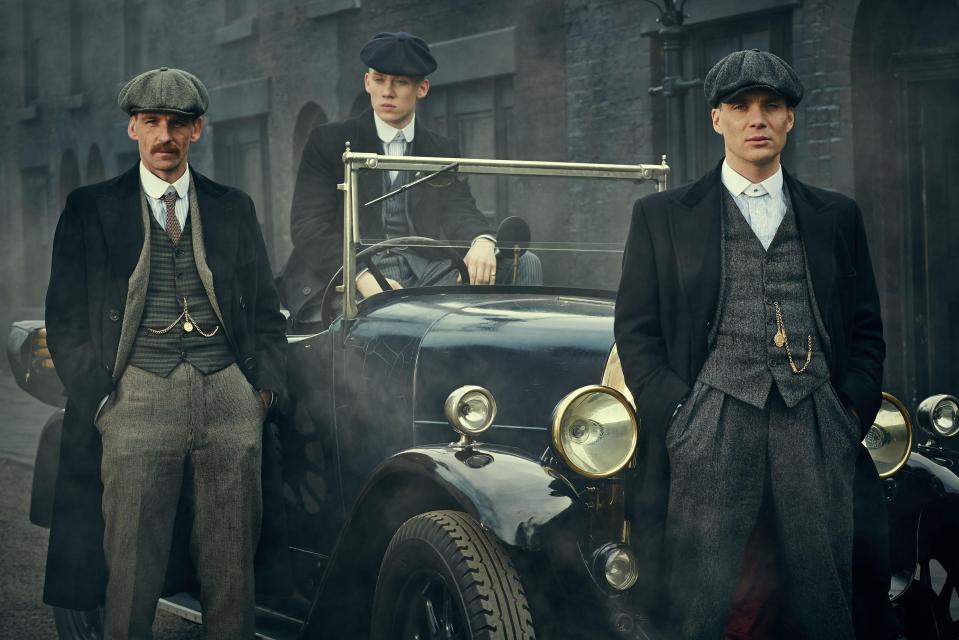 Three men in period attire standing by a vintage car.