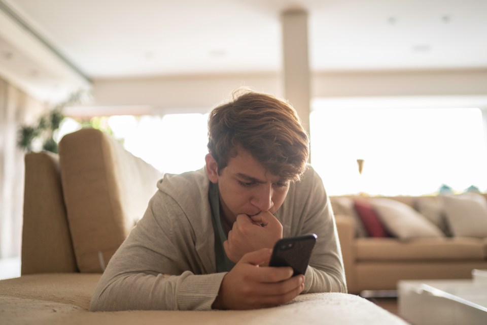 Teenage boy lying on a couch, looking at his smartphone with a serious expression.