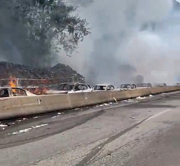 Aftermath of an ethanol tanker explosion, showing burning cars and trucks behind a concrete barrier.