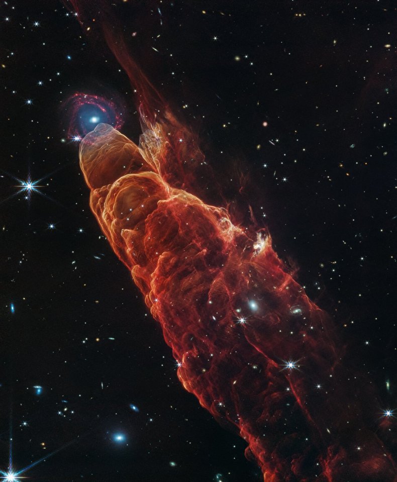 The NASA/ESA/CSA James Webb Space Telescope observed Herbig-Haro 49/50, an outflow from a nearby still-forming star, in high-resolution near- and mid-infrared light with the NIRCam and MIRI instruments. The intricate features of the outflow, represented in reddish-orange color, provide detailed clues about how young stars form and how their jet activity affects the environment around them. A chance alignment in this direction of the sky provides a beautiful juxtaposition of this nearby Herbig-Haro object (located within our Milky Way) with a more distant, face-on spiral galaxy in the background. Protostars are young stars in the process of formation that generally launch narrow jets of material. These jets move through the surrounding environment, in some cases extending to large distances away from the protostar. Like the water wake generated by a speeding boat, the arcs in this image are created by the fast-moving jet slamming into surrounding dust and gas. This ambient material is compressed and heats up, then cools by emitting light at visible and infrared wavelengths. In particular, the infrared light captured here by Webb highlights molecular hydrogen and carbon monoxide. The galaxy that appears by happenstance at the tip of Herbig-Haro 49/50 is a much more distant spiral galaxy. It has a prominent central bulge represented in blue that shows the location of older stars. It also displays hints of side lobes, suggesting that this could be a barred-spiral galaxy. Reddish clumps within the spiral arms show the locations of warm dust and groups of forming stars. In the image background there are many more galaxies at further distances, including galaxies that shine through the diffuse infrared glow of the nearby Herbig-Haro object. [Image description: Angled from the upper left corner to the lower right corner of the image is a conical shaped orange-red cloud known at Herbig-Haro 49/50. This feature takes up about three-fourths of the length of this angle. T