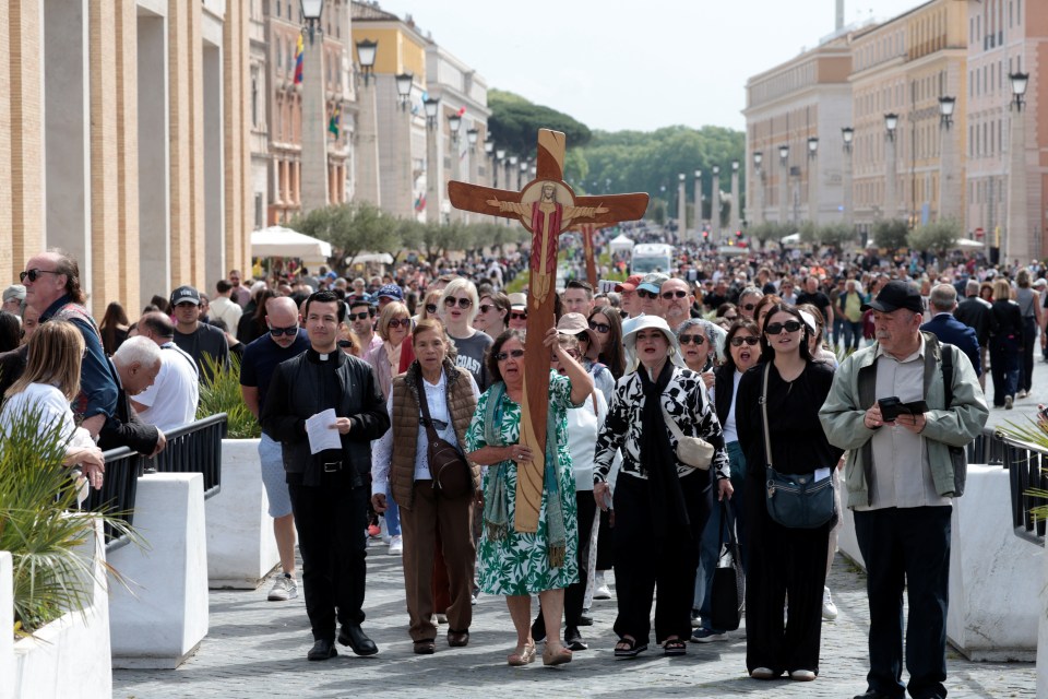 A woman carries a large cross as a crowd walks by after the death of Pope Francis was announced.