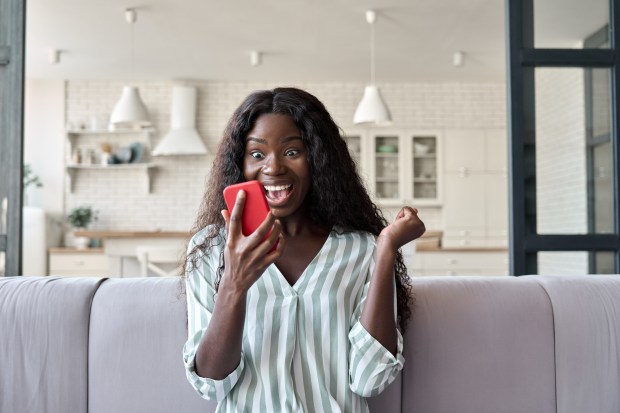 A young Black woman sits on a couch, excitedly looking at her phone.