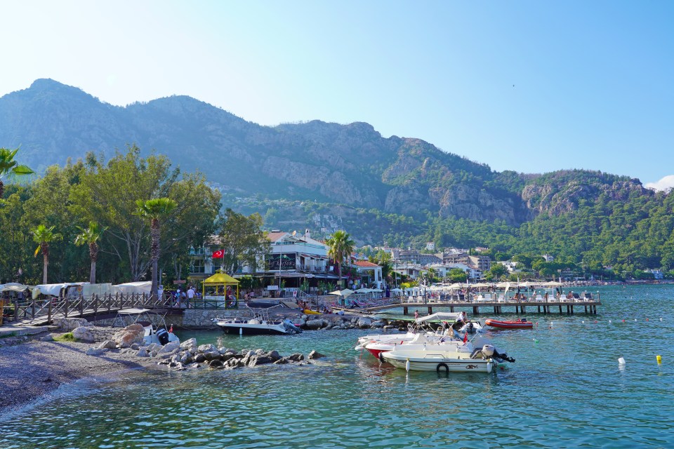 Aerial view of Turunç town in Marmaris, Turkey, showing boats moored at a pier, buildings, and mountains in the background.