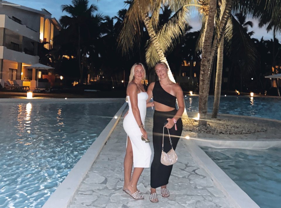 Two women in evening wear stand by a resort pool at night.