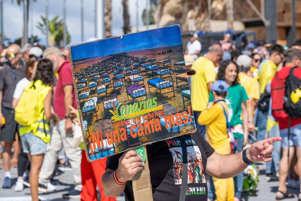 Protestor holding sign depicting hotel beds on a beach; text on sign reads "Canarias tiene un limite. Ni una cama mas!" (Canary Islands has a limit. Not one more bed!).