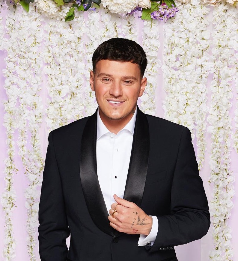 Man in tuxedo in front of floral backdrop.