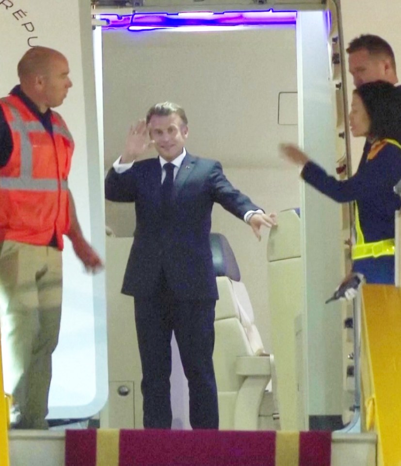 President Emmanuel Macron exiting a plane, waving, with staff members nearby.