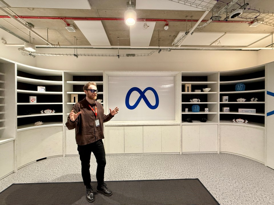 Man in glasses gesturing in front of a Meta logo displayed on a wall with shelves.