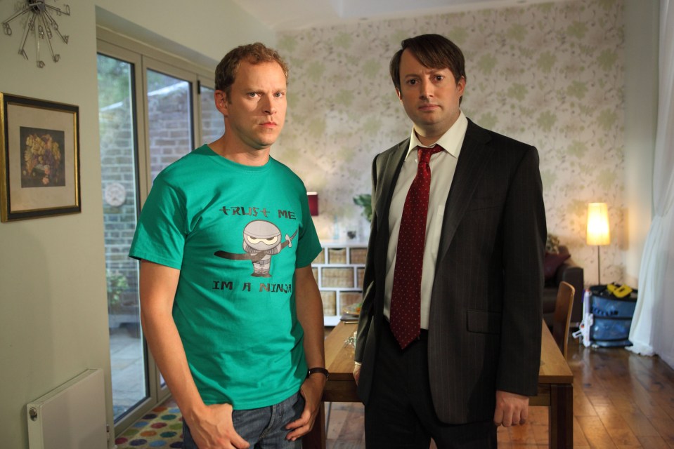 Two men standing in a living room; one wears a green t-shirt with a ninja illustration, the other a suit and tie.