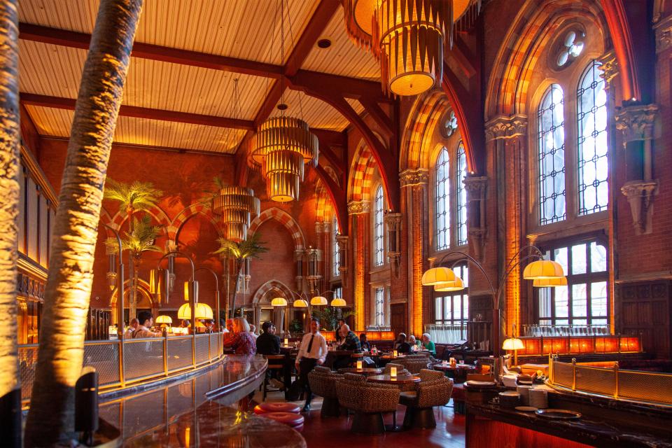 Interior view of a bar and restaurant with high ceilings, arched windows, and ornate lighting.