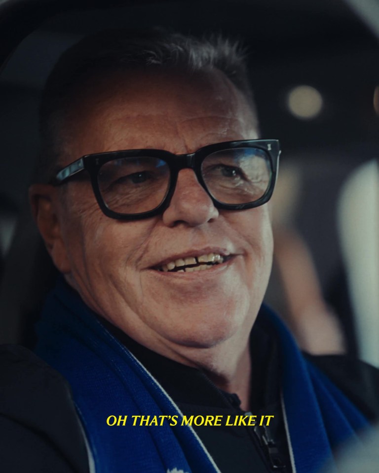Close-up of a man wearing glasses and a blue scarf, smiling and saying "Oh that's more like it."