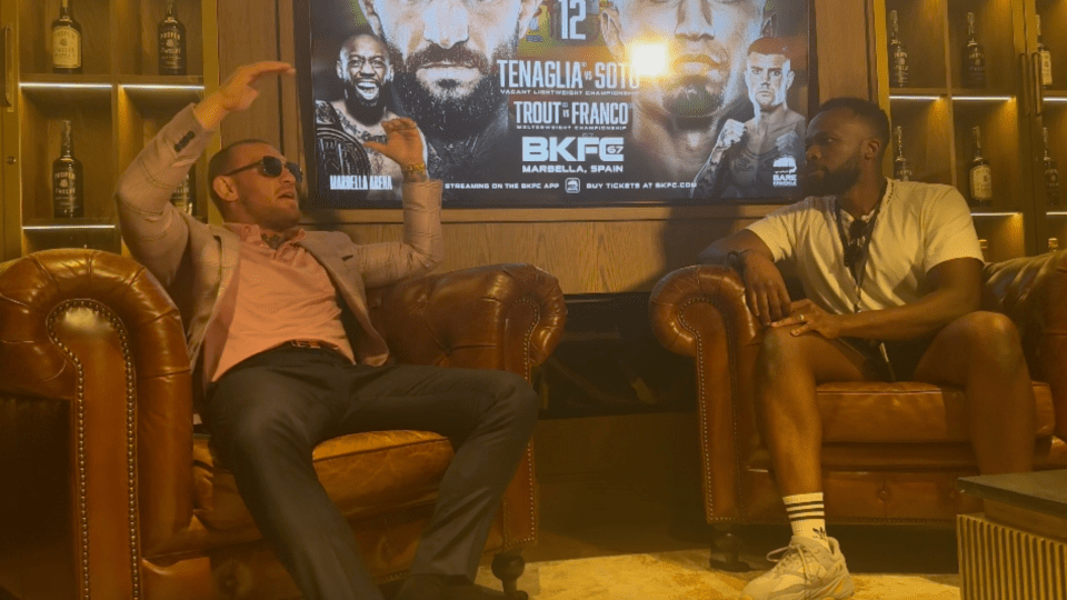 Two men sitting on leather chairs, discussing a BKFC fight poster.