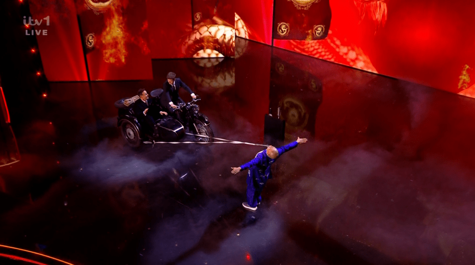 Screenshot of a man being pulled by a motorcycle sidecar on a stage.