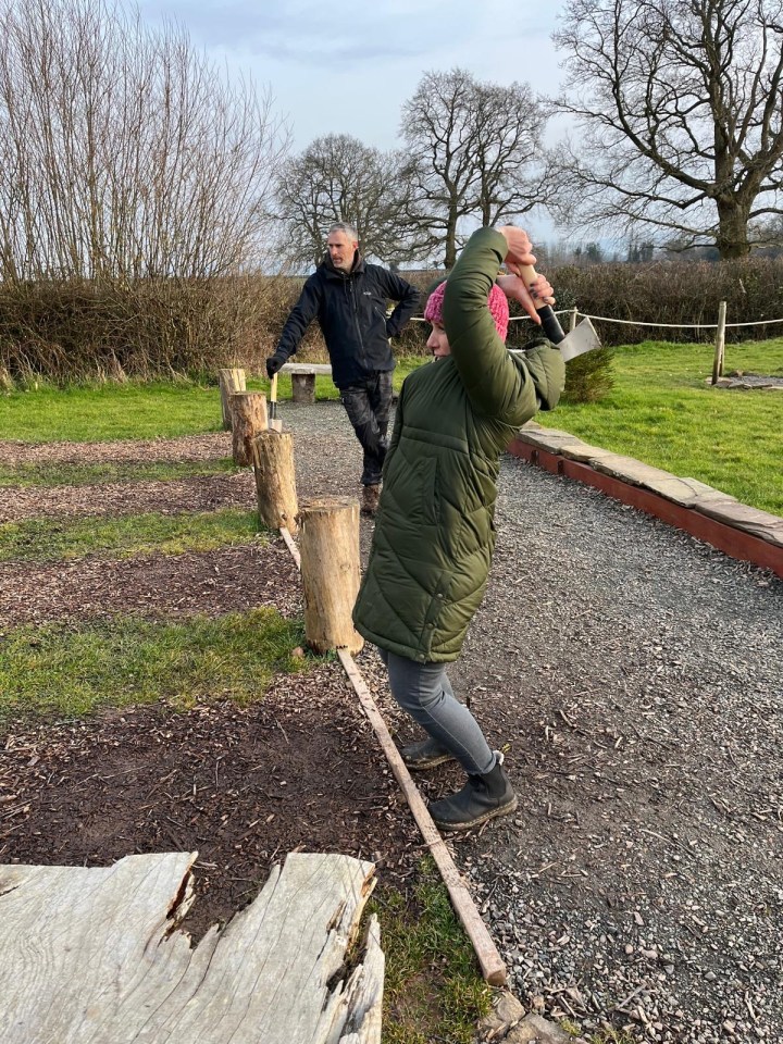 Woman throwing axe at wooden post, man watching.