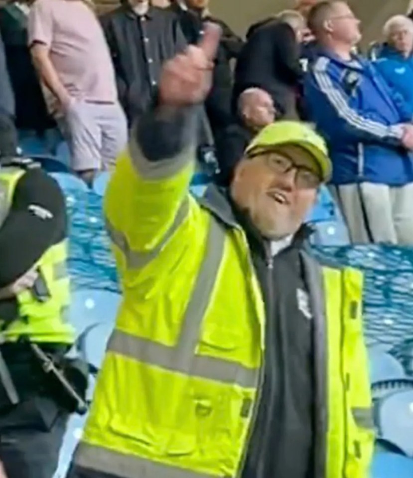 A man in a bright yellow safety jacket leads a crowd in a stadium.