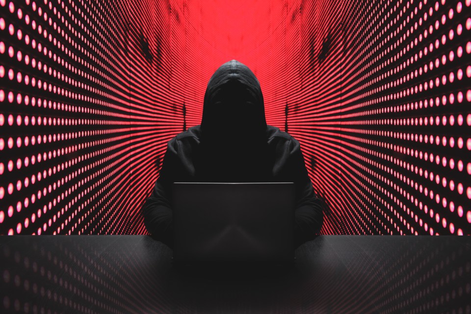 Hooded figure using a laptop in front of a red light wall.