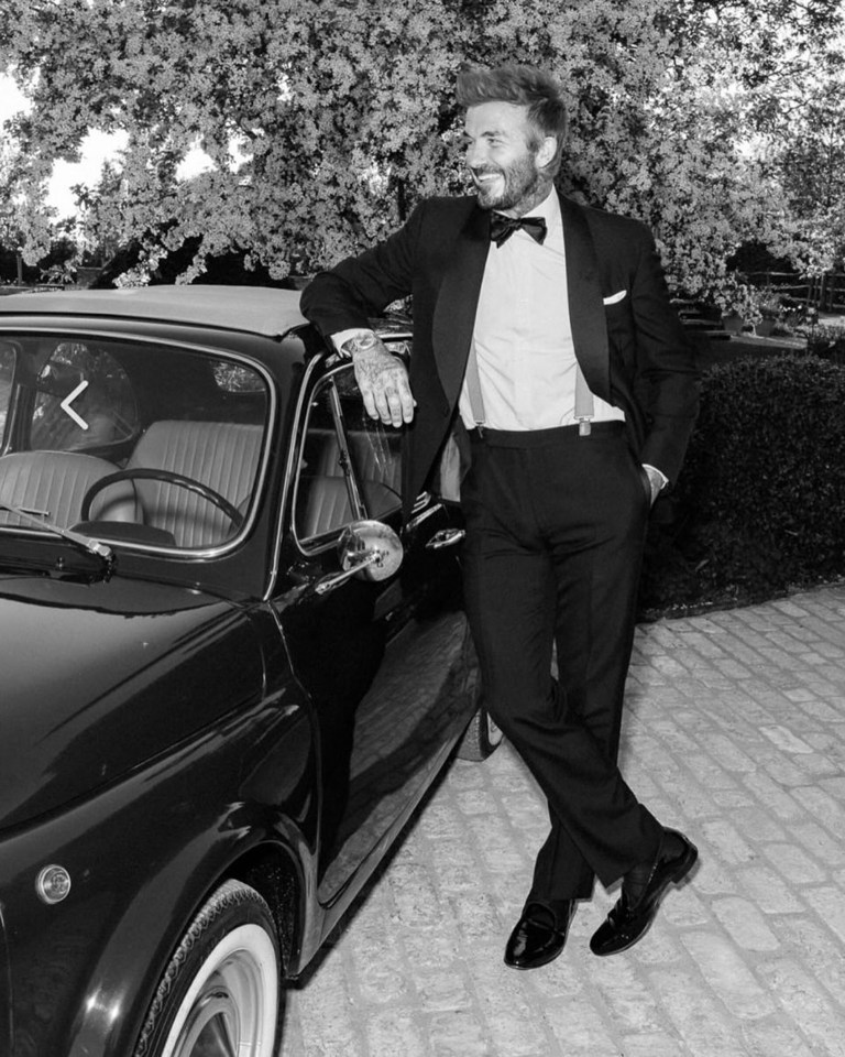 Black and white photo of David Beckham in a tuxedo leaning against a classic car.