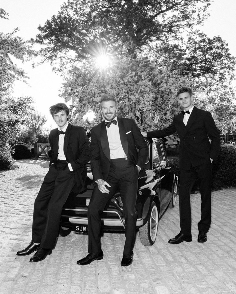 Black and white photo of David Beckham with two of his sons, all in tuxedos, posing with a classic car.