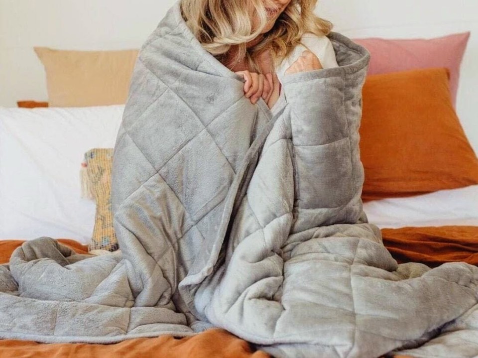 Person sitting on a bed wrapped in a gray weighted blanket.