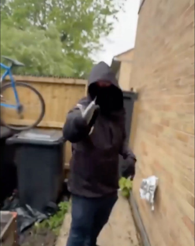 A hooded figure making a hand gesture in an alleyway.