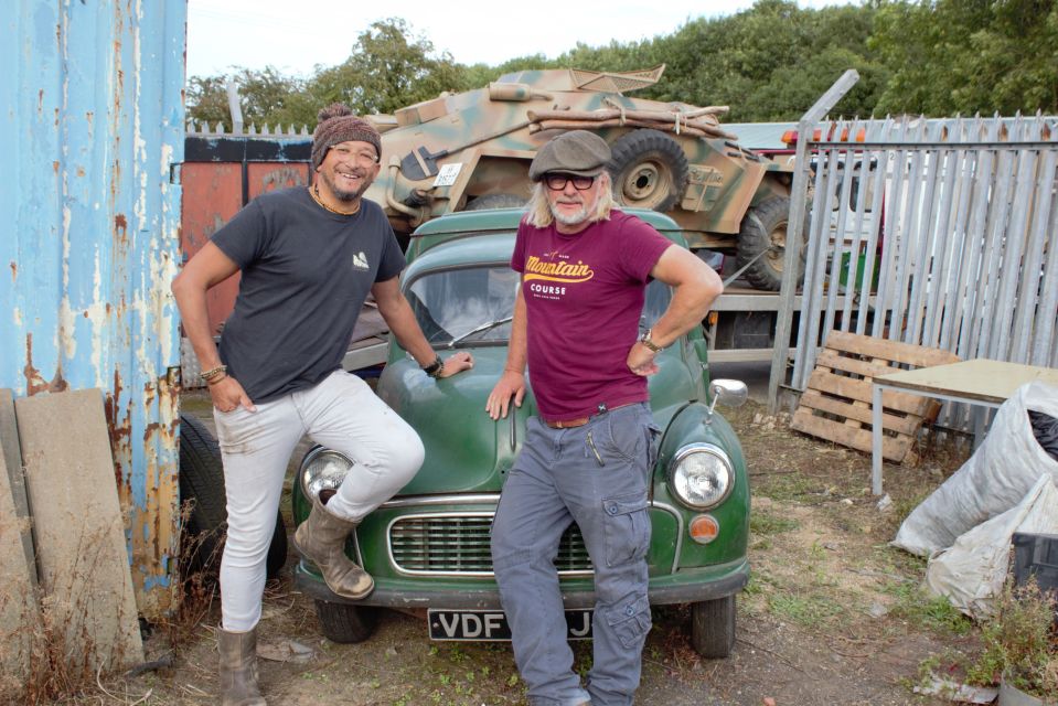 Two men standing by a vintage car in a junkyard.