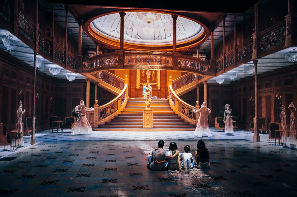 A family sits on the floor facing a digital recreation of the Titanic's Grand Staircase, with translucent figures of passengers dancing and mingling.