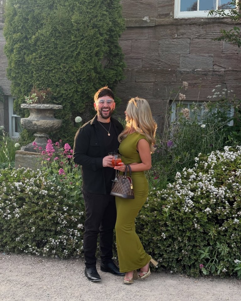 A man and woman smiling, holding drinks, outside a stone building.