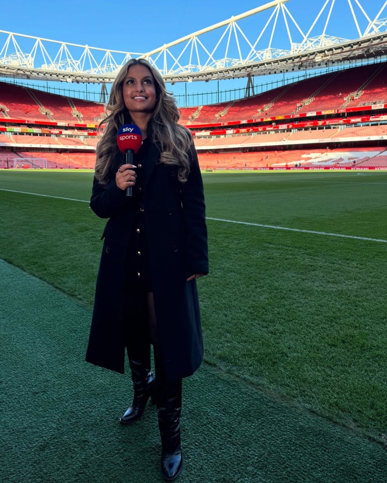 Woman in a black coat holding a Sky Sports microphone in front of a soccer stadium.