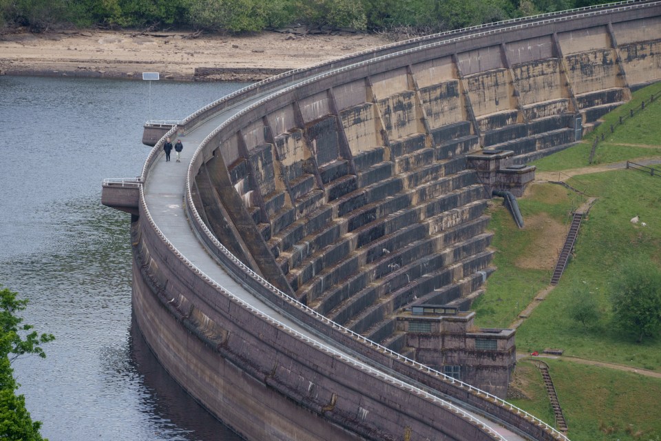 Two people walk along the top of a dam overlooking a reservoir.