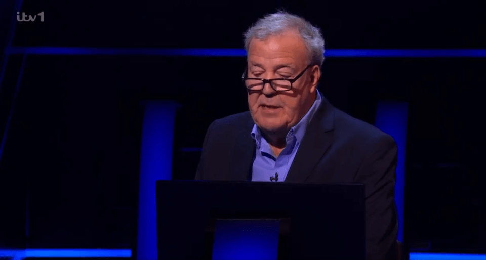 A man in a suit and glasses sits at a podium, appearing on a game show.