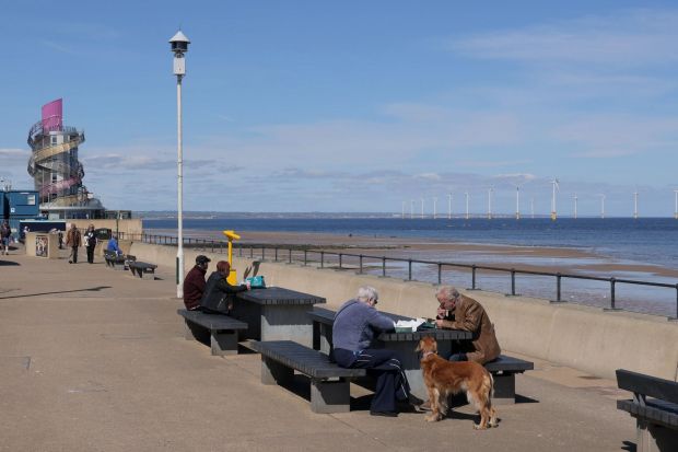 An image collage containing 1 images, Image 1 shows People sitting at picnic tables by the sea, with wind turbines visible in the distance