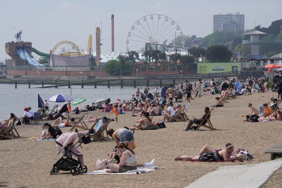 Crowded beach with people sunbathing and enjoying warm weather; amusement park in the background.